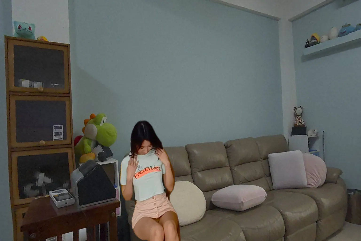 Asian woman wearing a light green T-shirt and a pink mini skirt, sitting on a sofa and gently pulling on her shirt while looking downward. Plush toys and cushions are visible in the background.